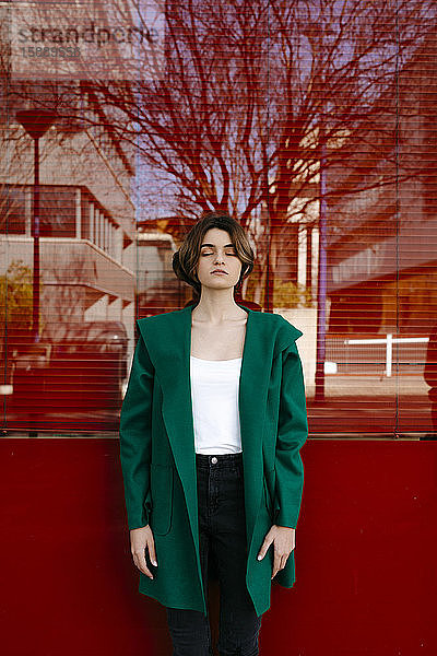 Junge Frau mit geschlossenen Augen trägt grünen Mantel vor einer Glasscheibe