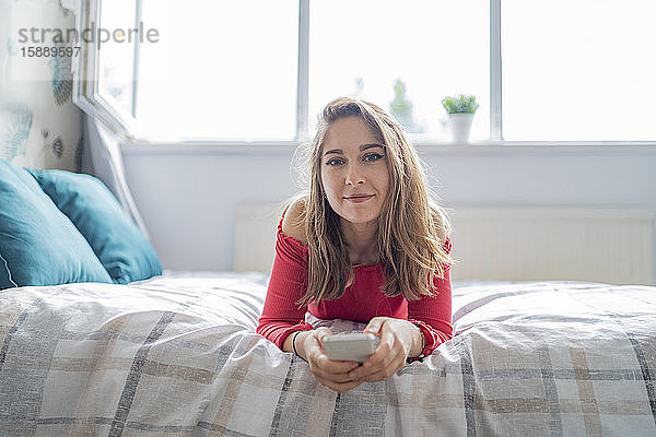 Porträt einer jungen Frau  die zu Hause auf dem Bett liegt und ein Handy hält