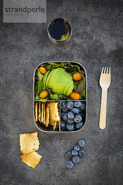 Schale mit Salatdressing und Brotdose mit Avocadoscheiben  gelben Tomaten  Crackern  Blaubeeren und grünem Salat