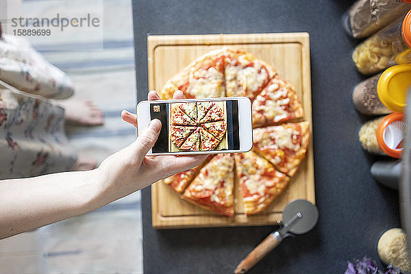 Ausschnittansicht einer Frau  die eine selbstgemachte Pizza mit einem Smartphone fotografiert