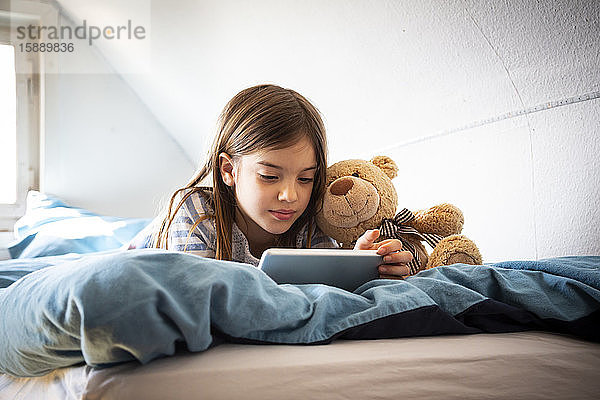Porträt eines auf dem Bett liegenden Mädchens mit Teddybär auf digitalem Tablett