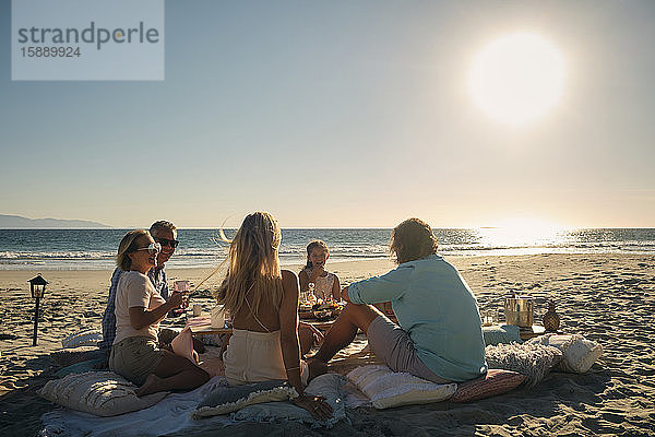 Ältere männliche und weibliche Freunde mit einem Mädchen  die bei Sonnenuntergang am Strand ein Picknick gegen den Himmel genießen. Riviera Nayarit  Mexiko