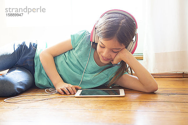 Mädchen liegt zu Hause auf dem Boden und benutzt Kopfhörer und digitales Tablett