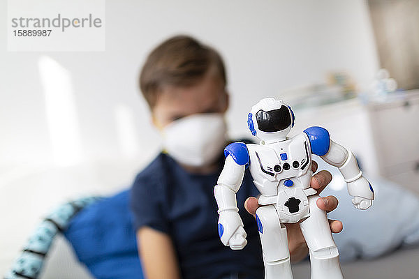 Junge mit Maske spielt während der Koronakrise mit seinem neuen  einzigen Freund  einem Roboter  in seinem Zimmer