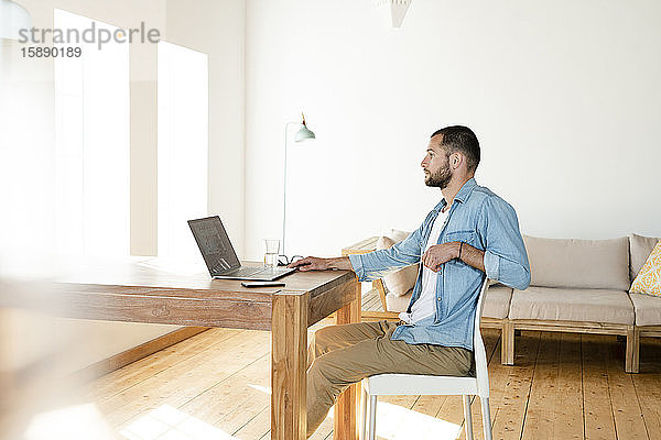Seriöser junger Mann arbeitet zu Hause am Laptop im Home-Office im modernen Wohnzimmer