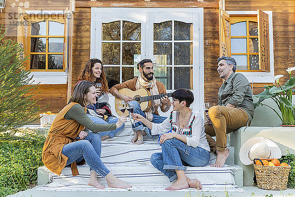 Freunde spielen Musik auf der Gitarre und trinken Wein vor einer Hütte auf dem Land