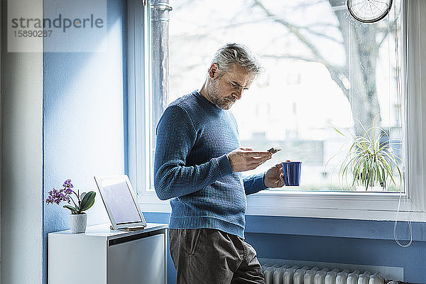 Reifer Mann steht mit Kaffeebecher im Wohnzimmer und schaut auf Smartphone