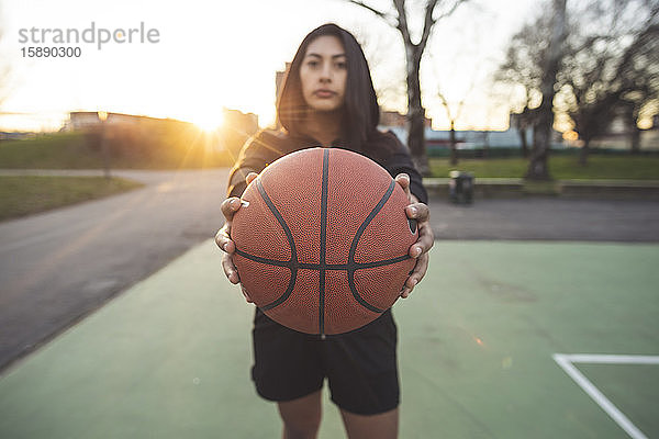 Junge sportliche Frau posiert bei Sonnenuntergang mit Basketball auf dem Platz