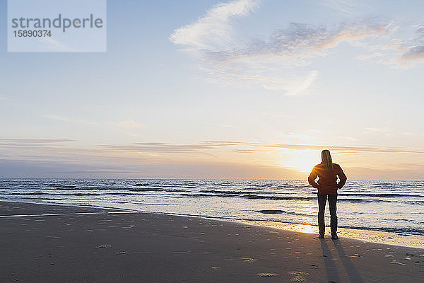 Frau in voller Länge an der Küste stehend  während sie bei Sonnenuntergang das Meer gegen den Himmel betrachtet  Nordseeküste  Flandern  Belgien