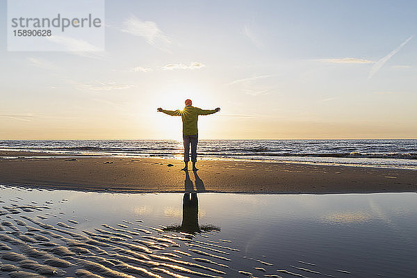 Rückansicht eines älteren Mannes in voller Länge mit ausgestreckten Armen  der bei Sonnenuntergang am Strand am Ufer steht  Nordseeküste  Flandern  Belgien