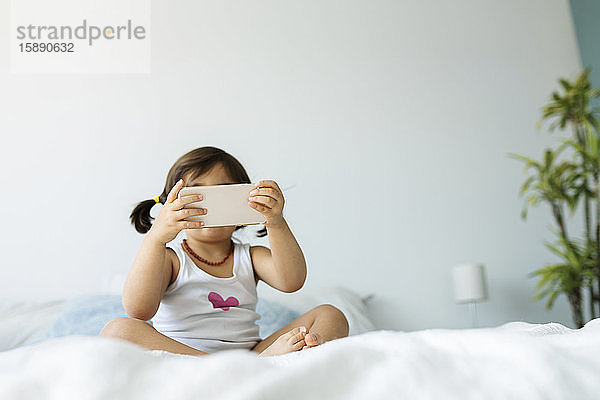 Kleines Mädchen sitzt in Unterwäsche auf dem Bett und benutzt ein Smartphone