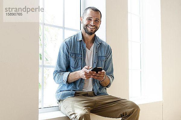 Glücklicher junger Mann zu Hause  der sein Smartphone in der Hand hält und auf der Fensterbank sitzt