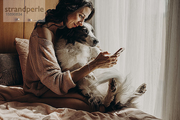 Lächelnde Frau sitzt mit ihrem Hund auf dem Bett und nimmt Selfie mit Smartphone