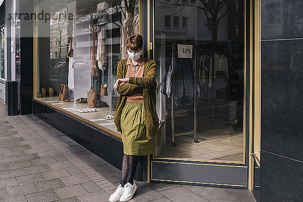 Frau mit Maske steht vor ihrem geschlossenen Bekleidungsgeschäft