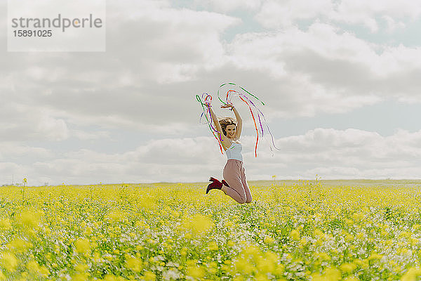 Glückliche junge Frau springt im Frühling mit bunten Bändern auf einer Blumenwiese