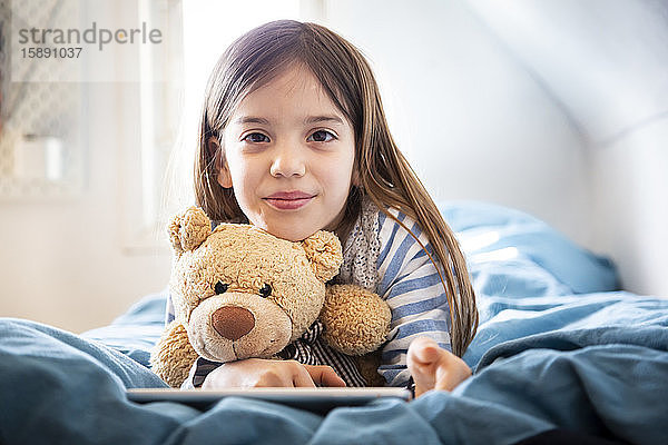 Porträt eines lächelnden  auf dem Bett liegenden Mädchens mit Teddybär und digitalem Tablett