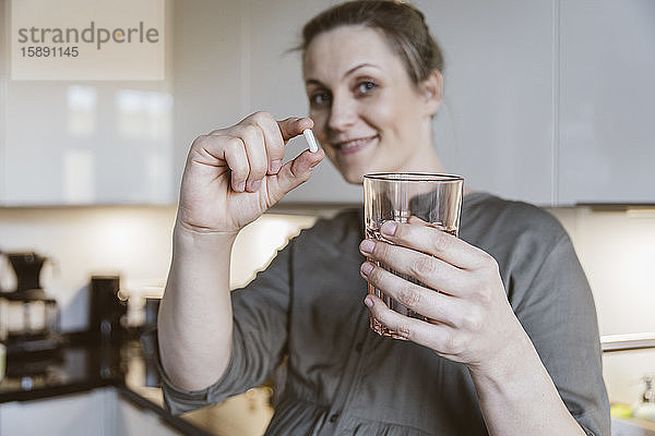 Porträt einer Frau  die zu Hause in der Küche eine Pille und ein Glas Wasser in der Hand hält