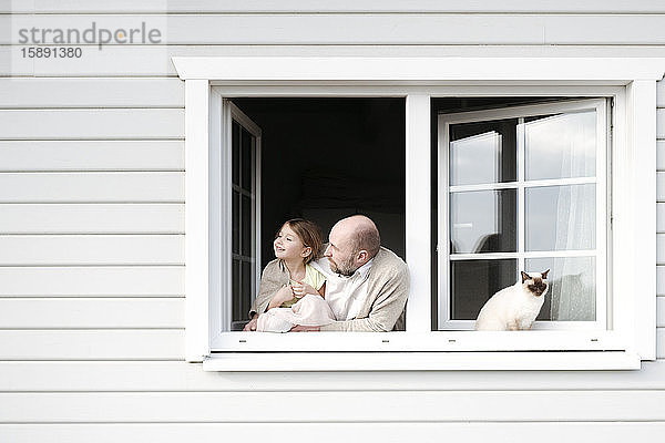 Reifer Mann mit lächelnder Tochter und Katze  die sich aus dem Fenster seines Hauses lehnt