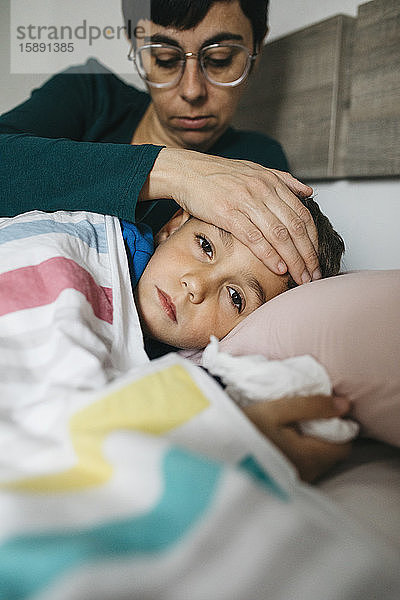 Porträt eines kranken Jungen  der im Bett liegt  während seine Mutter seine Stirn berührt