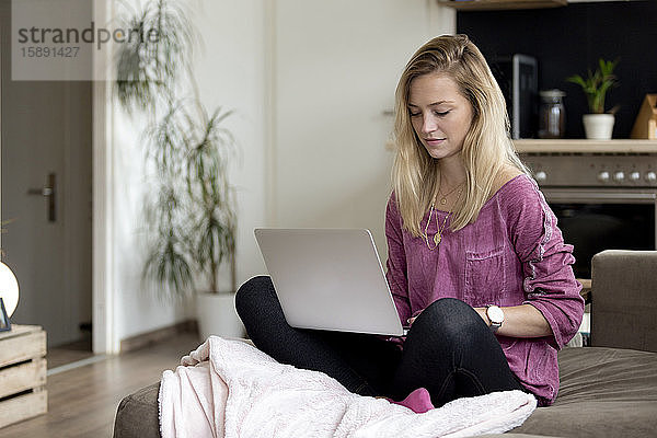 Porträt einer blonden jungen Frau  die zu Hause auf einer Couch sitzt und einen Laptop benutzt