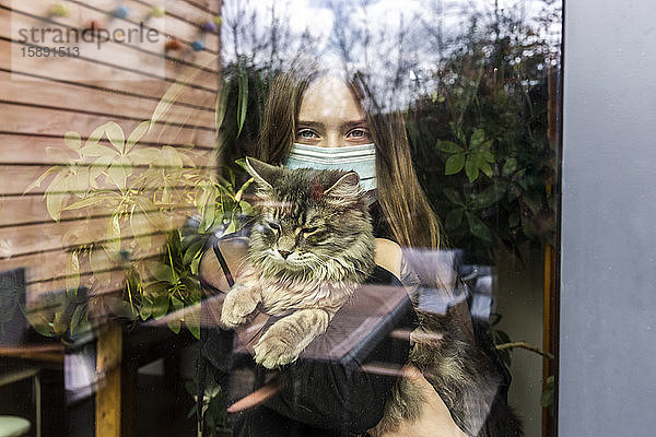 Porträt eines Mädchens mit chirurgischer Maske und Katze hinter Fensterscheibe