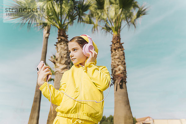 Porträt eines kleinen Mädchens mit gelber Kapuzenjacke  das mit Kopfhörer und Smartphone Musik hört