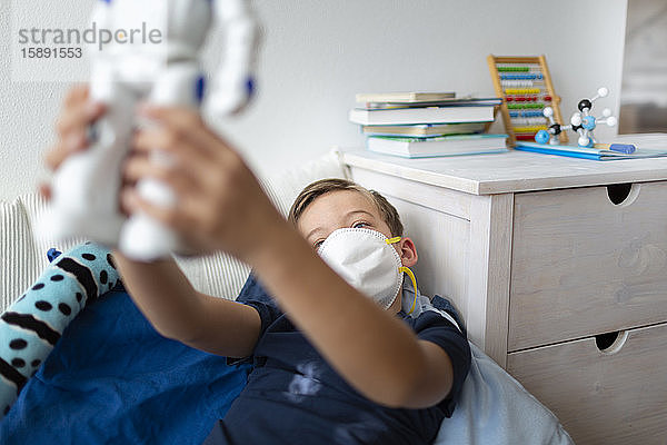 Junge mit Maske spielt während der Koronakrise mit seinem neuen  einzigen Freund  einem Roboter  in seinem Zimmer