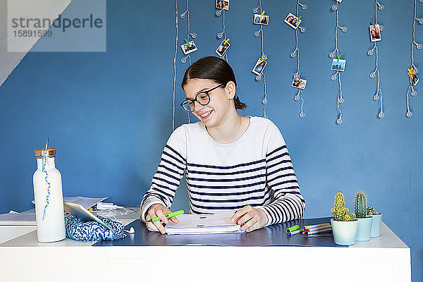 Porträt eines lächelnden Mädchens  das zu Hause am Schreibtisch sitzt und auf ein digitales Tablet schaut