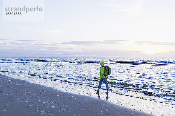 Pensionierter älterer Mann in voller Länge mit Rucksack  der bei Sonnenuntergang am Strand am Ufer spazieren geht  Nordseeküste  Flandern  Belgien