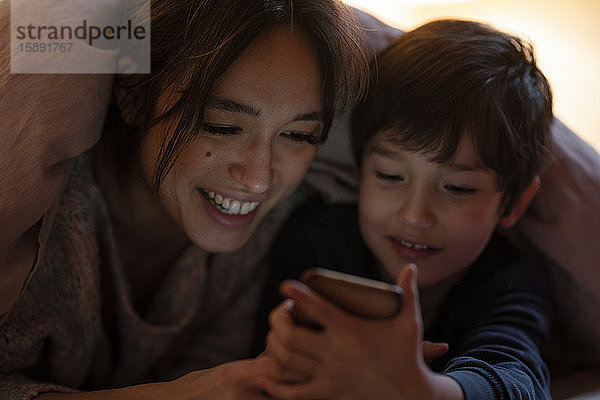 Mutter und kleiner Sohn liegen zusammen mit einer Decke zugedeckt auf dem Bett und sehen sich einen Film auf einem Smartphone an