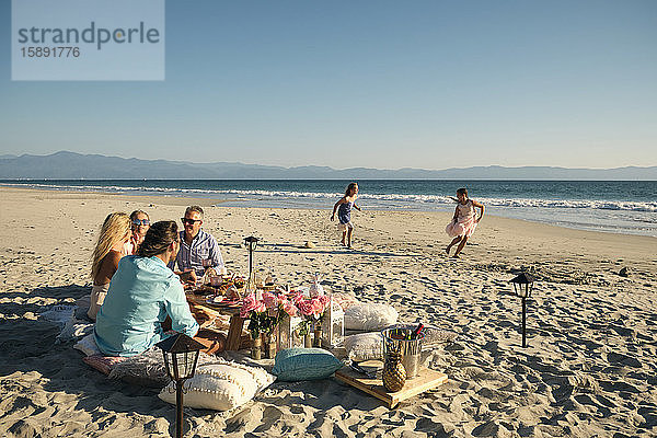 Ältere männliche und weibliche Freunde genießen das Picknick  während die Mädchen am Strand bei strahlendem Sonnenschein gegen den klaren Himmel spielen. Riviera Nayarit  Mexiko