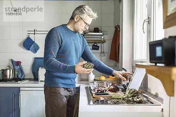 Reifer Mann bereitet Salat in seiner Küche mit digitalem Tablett zu