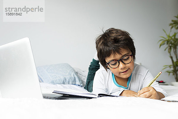Porträt eines kleinen Jungen  der mit einem Laptop auf dem Bett liegt und Hausaufgaben macht