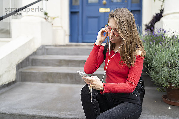Junge Frau sitzt am Hauseingang mit Kopfhörern und Smartphone