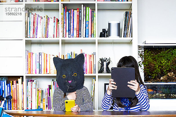Zwei Mädchen sitzen am Tisch und verstecken sich hinter einem digitalen Tablett und einer Katzenmaske