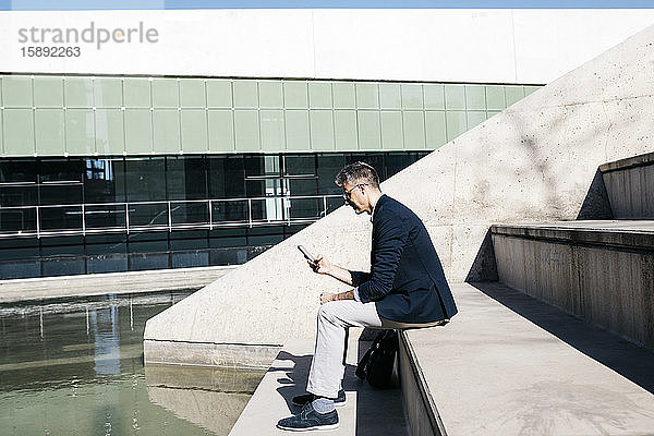 Grauhaariger Geschäftsmann sitzt auf einer Treppe an einem Pool und benutzt sein Handy