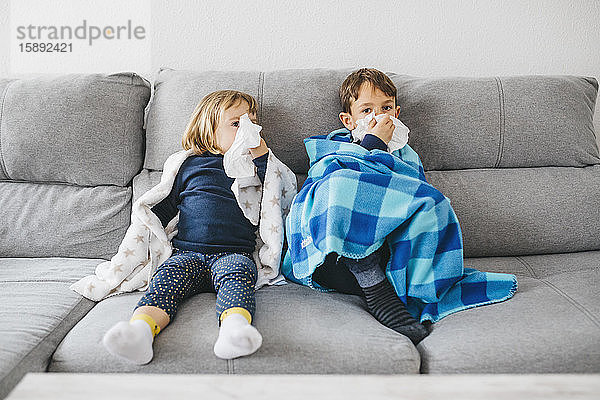 Kleines Mädchen und ihr Bruder sitzen zu Hause nebeneinander auf der Couch und putzen sich die Nase