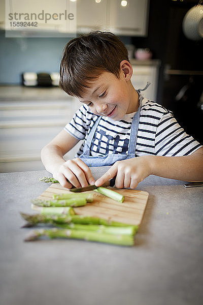 Porträt eines lächelnden kleinen Jungen  der in der Küche grünen Spargel schneidet