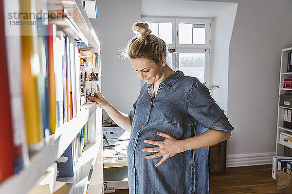 Schwangere Frau steht zu Hause am Bücherregal