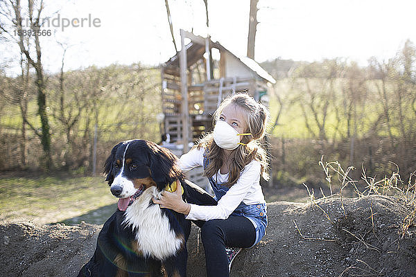 Mädchen mit Gesichtsmaske  sitzt im Garten und spielt mit ihrem Hund