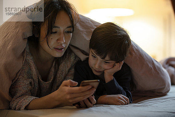 Mutter und kleiner Sohn liegen zusammen mit einer Decke zugedeckt auf dem Bett und sehen sich einen Film auf einem Smartphone an