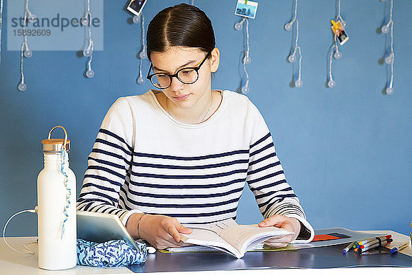 Porträt eines Mädchens  das mit einem Schulbuch am Schreibtisch sitzt und auf ein digitales Tablett schaut