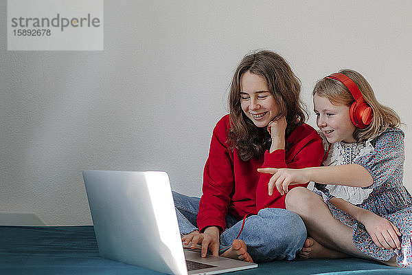 Porträt von zwei Schwestern  die zusammen auf dem Bett sitzen und auf einen Laptop schauen