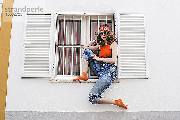 Porträt einer jungen Frau mit Stirnband und Sonnenbrille  die auf einem Fensterbrett im Freien sitzt