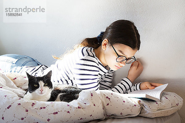 Mädchen liegt mit Katze auf dem Bett und liest ein Buch