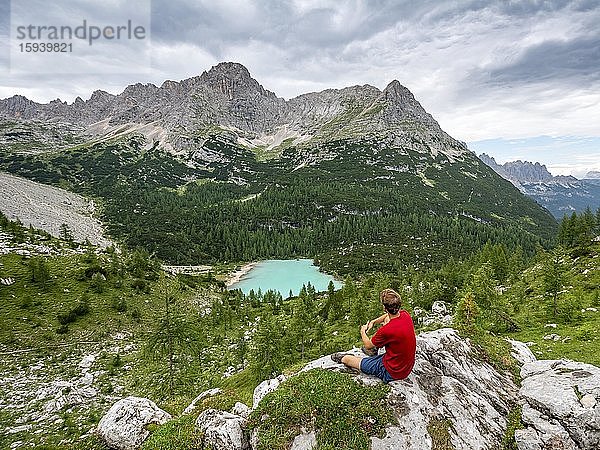 Junger Mann  Wanderer sitzt auf Felsen und blickt auf türkisgrünen Sorapissee und Berglandschaft  Dolomiten  Belluno  Italien  Europa