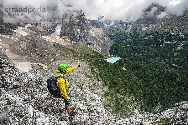 Junger Mann deutet in die Ferne  Bergsteiger an einem Klettersteig  Via Ferrata Vandelli  Blick auf Lago di Sorapis  Sorapiss Umrundung  Berge mit tiefhängenden Wolken  Dolomiten  Belluno  Italien  Europa