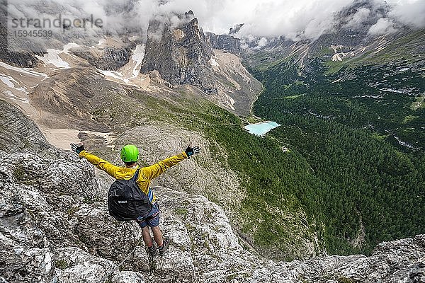 Junger Mann streckt Hände in die Luft  Bergsteiger an einem Klettersteig  Via Ferrata Vandelli  Blick auf Lago di Sorapis  Sorapiss Umrundung  Berge mit tiefhängenden Wolken  Dolomiten  Belluno  Italien  Europa