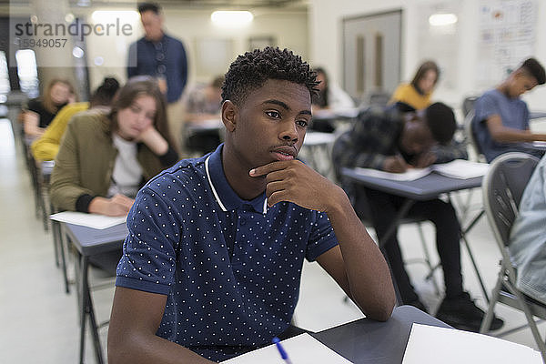 Nachdenklicher High-School-Junge nimmt Prüfung am Schreibtisch im Klassenzimmer ab