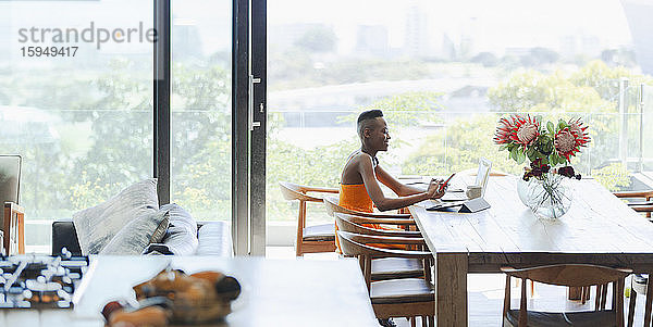 Junge Frau mit Smartphone und Laptop  die von zu Hause am Esstisch arbeitet